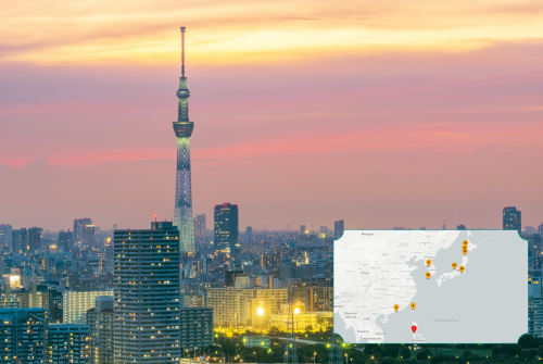 Stadtansicht von Tokio bei Sonnenuntergang mit dem beleuchteten Tokyo Skytree im Zentrum; im Vordergrund dichte Hochhäuser, im Hintergrund ein orange-rosa Himmel. Unten rechts ist eine kleine Karte von Japan eingeblendet, auf der mehrere markierte Punkte die Route der Kreuzfahrt darstellen.