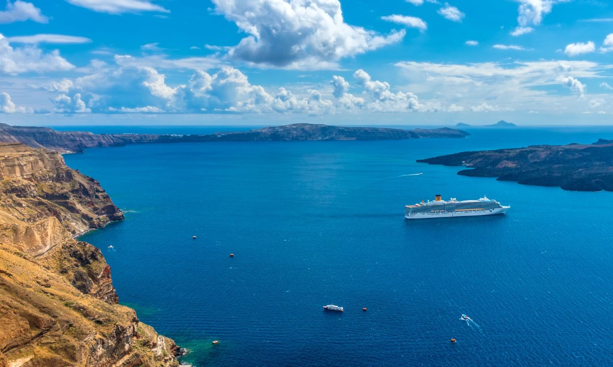 Großes Kreuzfahrtschiff in einer tiefblauen Meeresbucht bei Santorin, umgeben von steilen, braunen Felsküsten unter leicht bewölktem Himmel.