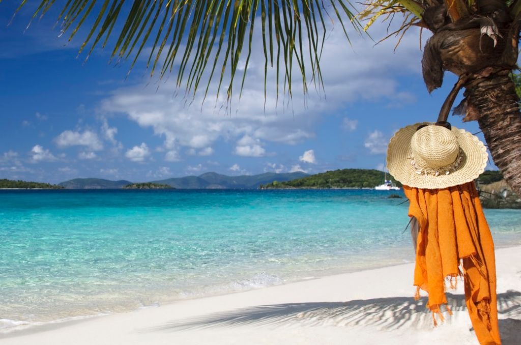 Tropischer Sandstrand mit türkisblauem Meer. Rechts hängt ein Strohhut mit orangefarbenem Tuch an einem Palmenstamm. Tropischer Sandstrand mit türkisblauem Meer. Rechts hängt ein Strohhut mit orangefarbenem Tuch an einem Palmenstamm.